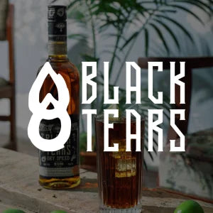 Eine Flasche Black Tears Single Malt Whiskey steht auf einem Holztisch, davor ein mit Whiskey und Eis gefülltes Glas, im unscharfen Hintergrund einige grüne Limetten und Pflanzen.