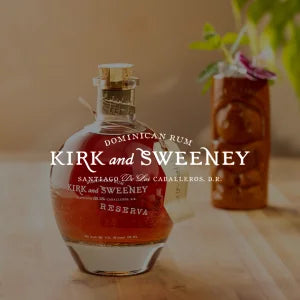 Produktfoto einer dunkelgold-braunen Rum-Flasche mit dem Aufdruck ›Kirk and Sweeney Dominican Rum Reserva‹, stehend auf einer hellen Holzoberfläche mit unscharfem Hintergrund.